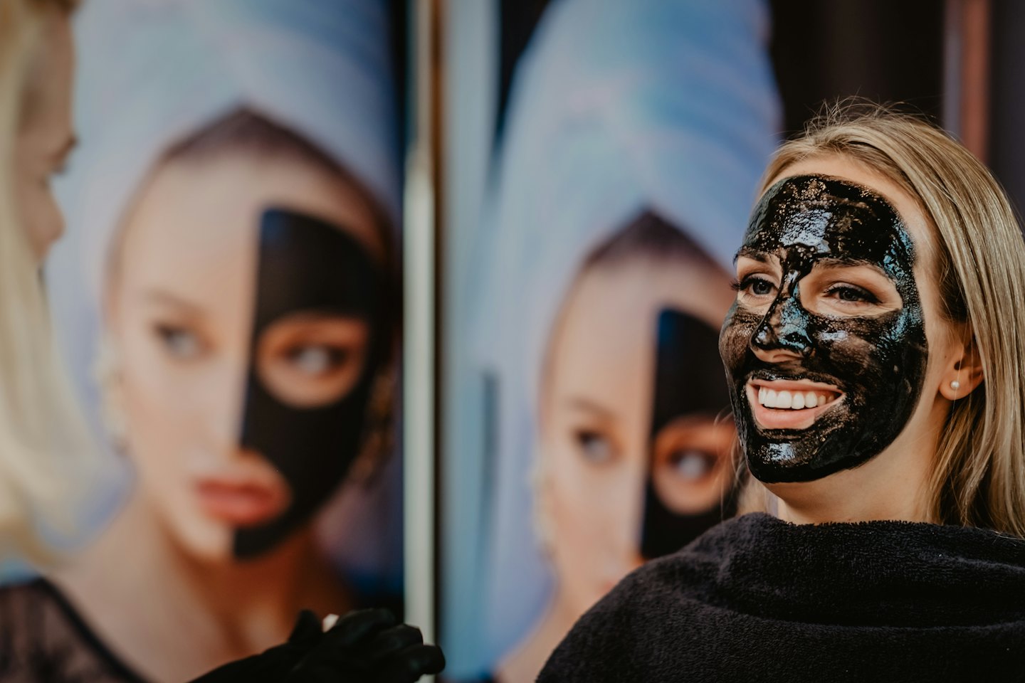 Vrouw lacht tijdens een gezichtsbehandeling met een houtskoolmasker.